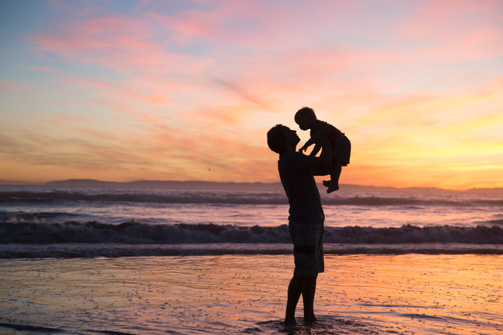 A man holding a small child in the air at the beach during sunset